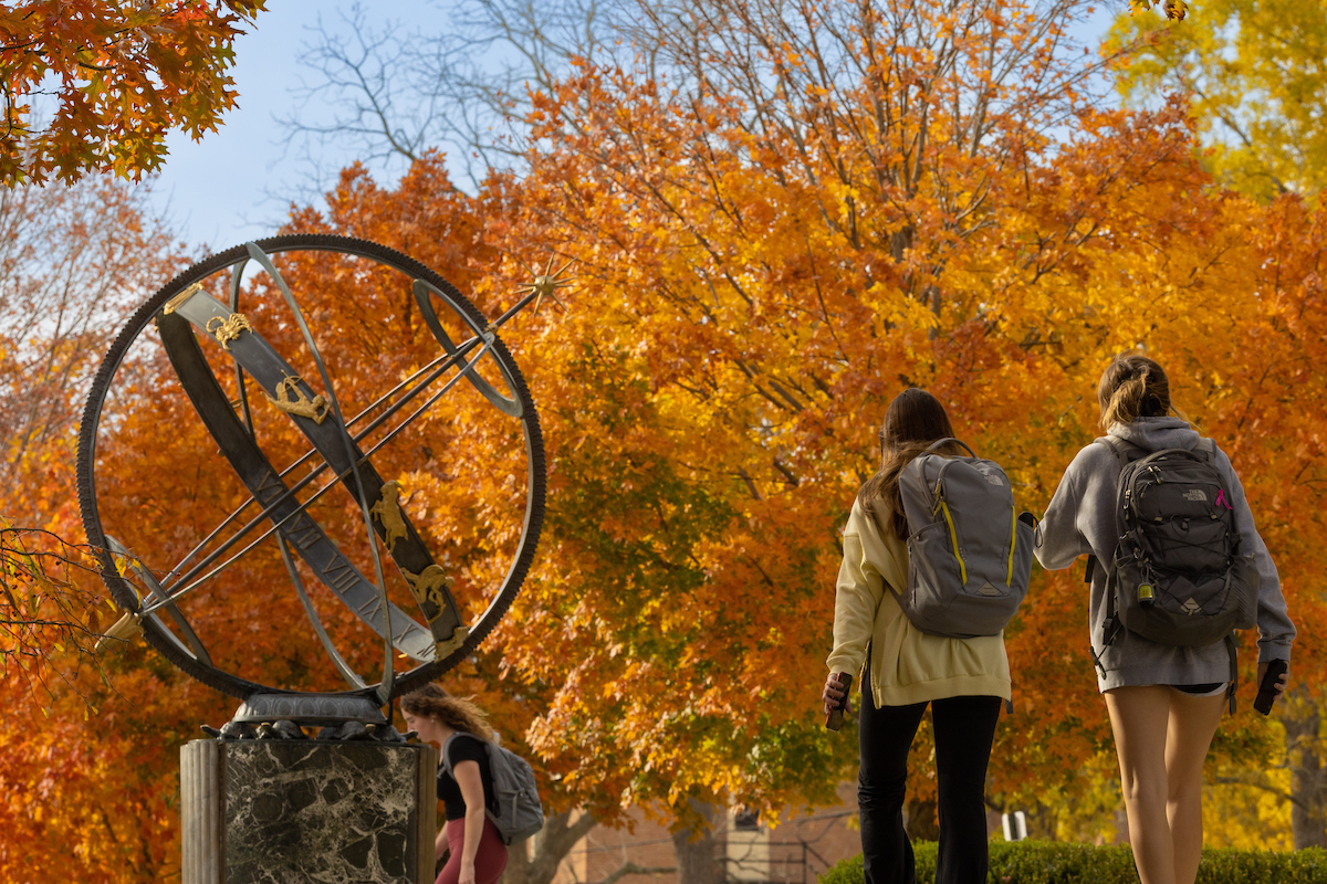 Two students are carrying their backpacks as they walk past the campus sundial. There are orange fall leaves in the background.