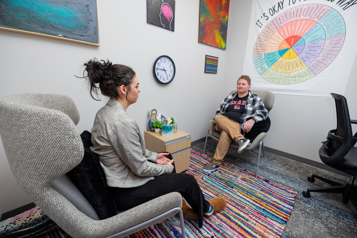 Two individuals sit facing one another in a counseling office with wellness resources surrounding them