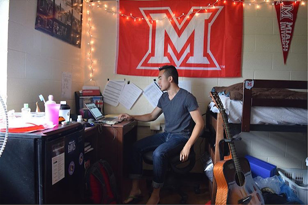 Miami student working at his desk in his residence hall room, a string of lights adorn a Miami flag on the wall