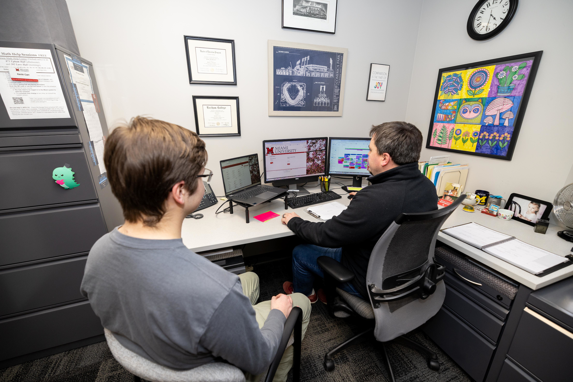 A professor works on a computer while a student looks on.