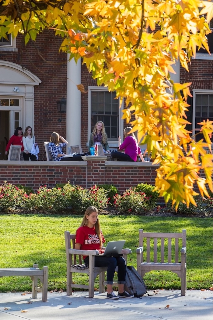 Several students hanging out on the patio of a dorm, and one female student sitting in a chair in front of the patio working on her laptop