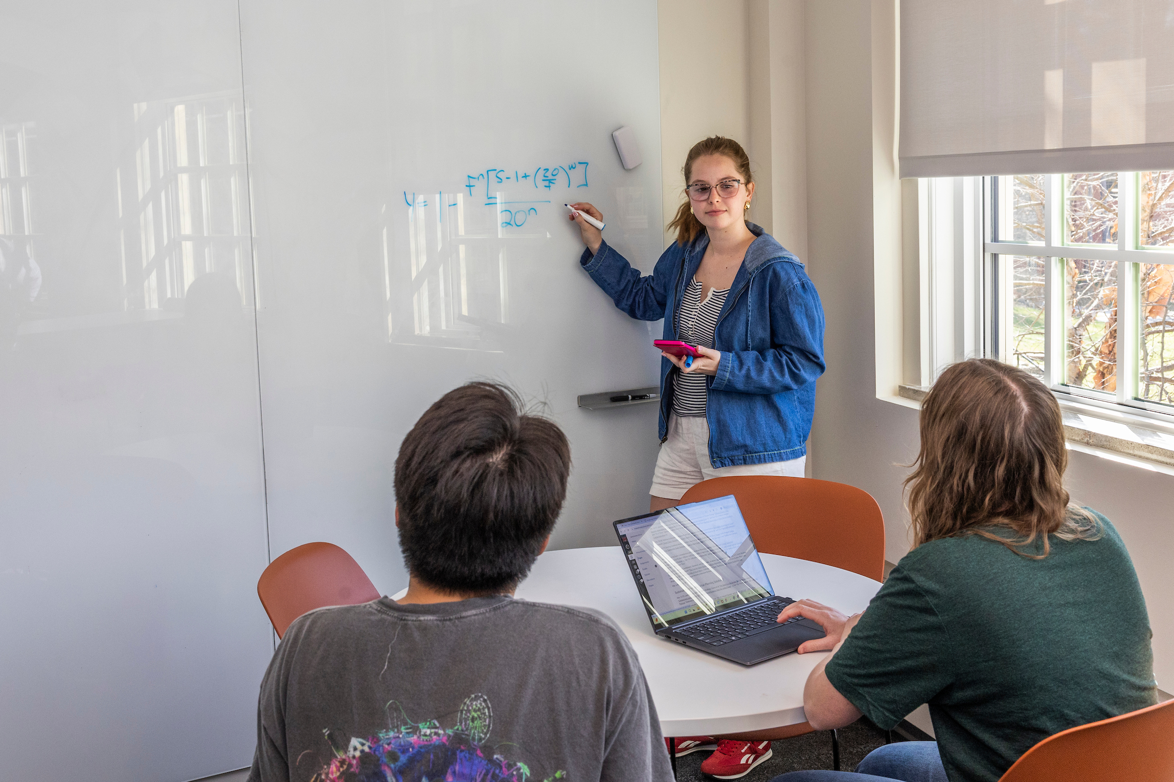 A student writes on a white board while two seated students look on.