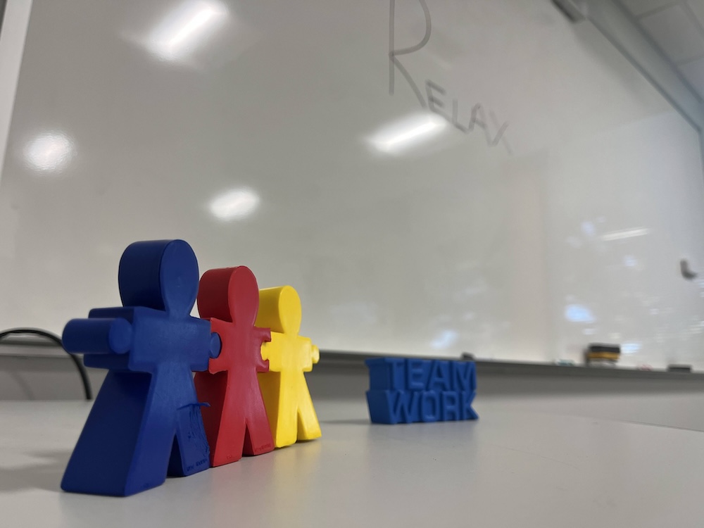 Stress squeeze toys in the shape of people and the word teaamwork sitting on a table with the word relax written on a whiteboard behind them
