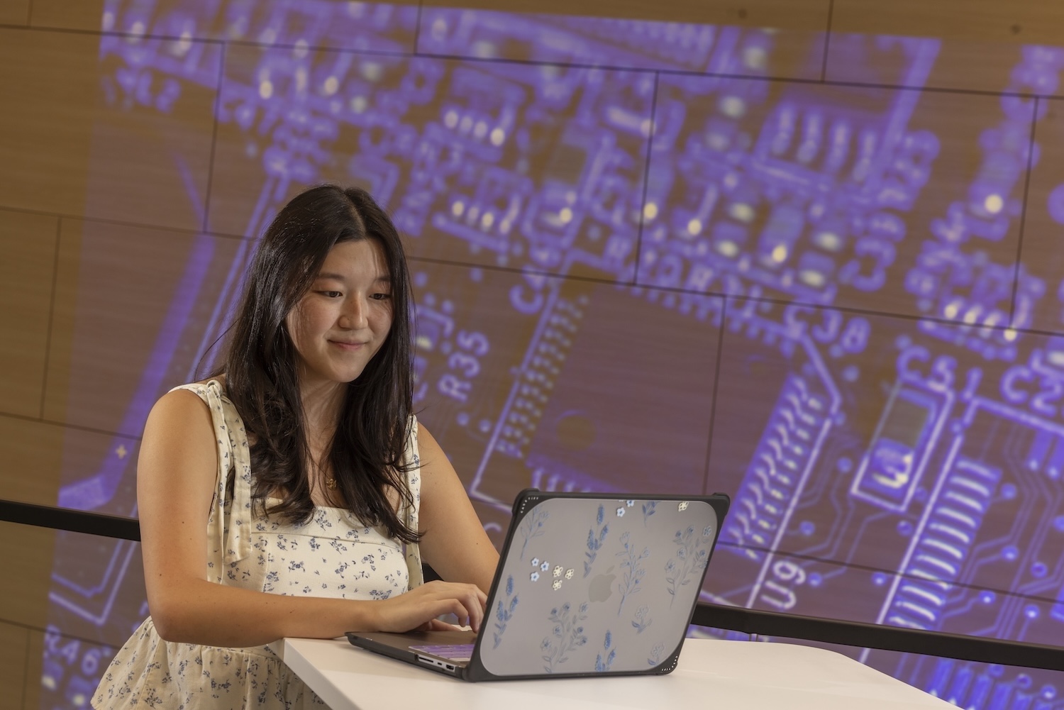 A student standing at a high-top table working on a laptop computer in front of a screen with a technology light show displaying